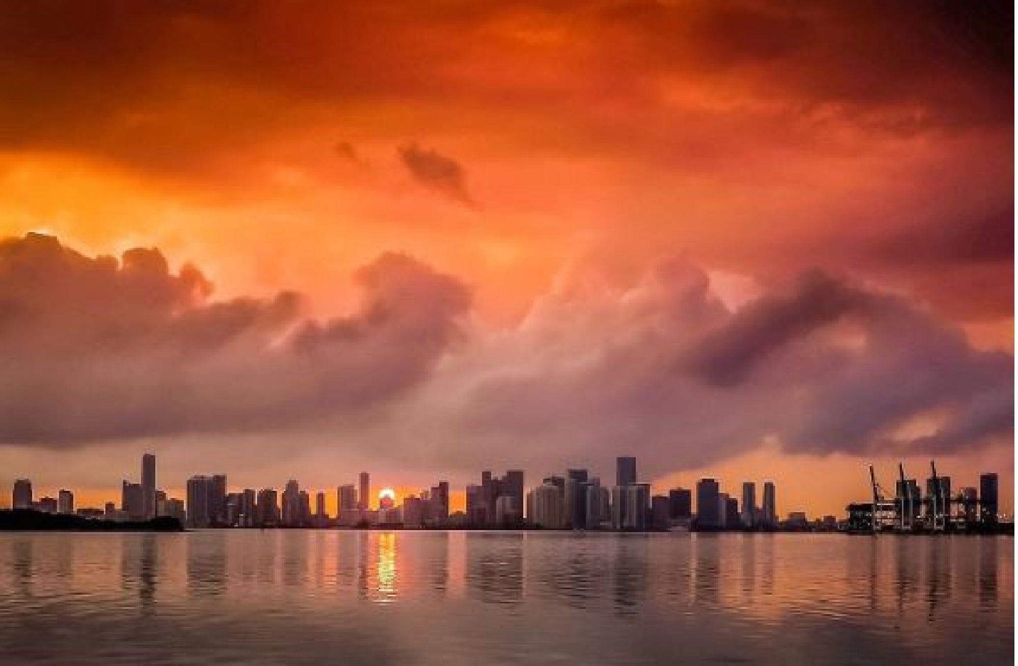 A city skyline at sunset with a dramatic orange and red sky, clouds, and reflections on calm water.