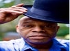 A person with a calm expression lifts a black bowler hat off their head. They are wearing a light blue shirt, and there is a blurred outdoor background.