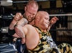 Two male wrestlers are engaged in a match; one is applying a hold from behind while the other wears a camouflage outfit with a skull design.