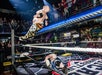 A wrestler in mid-air leaps off the ropes toward an opponent lying on the mat in a ring, surrounded by a crowd of spectators in an indoor venue.