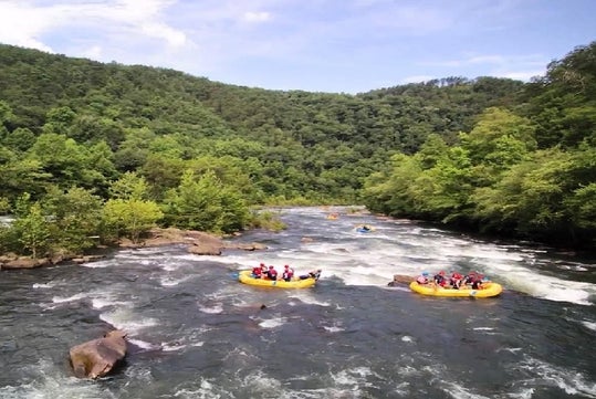 Two yellow rafts with people whitewater rafting on a river surrounded by green, forested hills under a partly cloudy sky.