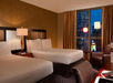 Hotel room with two double beds, a lamp, upholstered chair, and desk. View of city buildings and neon lights through the window at night.