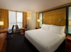 Hotel room with a large bed, two chairs, a small table, a desk with a chair, and a floor lamp by a window with curtains. The color scheme includes beige, gold, and brown tones.