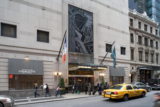 Urban building exterior with a central entrance, people walking, a yellow taxi on the street, and the flags of France and another country displayed beside the entry.