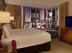 Hotel room with a king-size bed, a chair near a corner window view of a cityscape at night, a dresser with a TV, and a lamp on a side table.