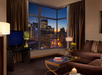 A cozy living room with a dark sofa, TV, and glass coffee table. Large windows reveal a city skyline at dusk, with glowing buildings and a darkening sky.