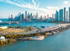 Aerial view of a cityscape with tall buildings, a winding bridge, and a coastline with blue waters and a clear sky.