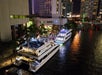 Two illuminated yachts docked by a city waterfront at night, with people visible on deck and tall buildings in the background.