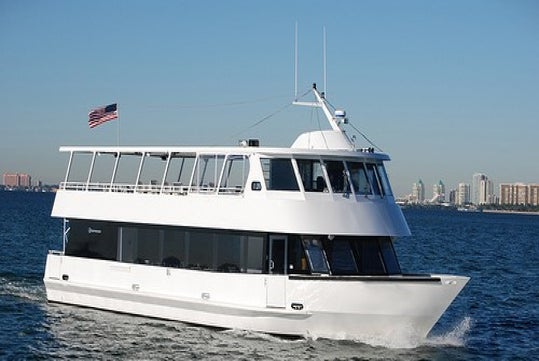 White double-deck boat on water with a U.S. flag at the stern, cityscape in the background under a clear blue sky.