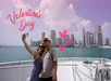 A couple takes a selfie on a boat in front of a city skyline under a pink sky with "Valentine's Day" and heart graphics.