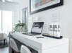A minimal desk with a laptop, two white stools, a plant, and two boxed water containers in a bright room.