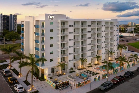 A modern multi-story apartment building with balconies, a pool area, and palm trees is seen at sunset, surrounded by parked cars and cityscape.