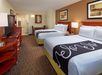 Hotel room with two double beds, a TV, a lamp, and a small table with chairs. The walls are decorated with framed art, and the carpet is brown.