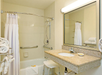 Bathroom with a shower-tub combo, white shower curtain, and a large mirror above a granite countertop sink. Various towels are neatly arranged, and a shower chair is placed inside the tub.