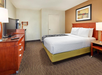 Hotel room with a double bed, wooden furniture, a TV, and a coffeemaker on a dresser. A framed abstract painting hangs above the bed. Neutral-toned walls and carpet.