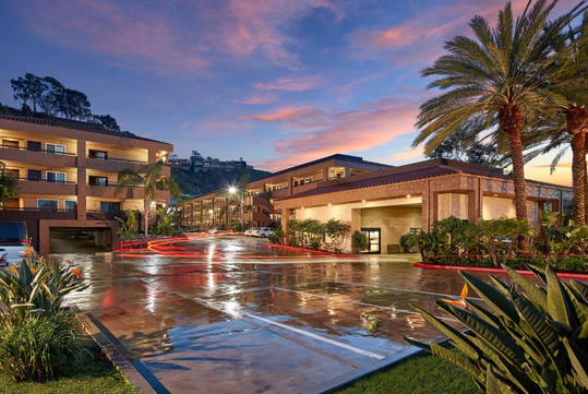 Hotel exterior at sunset with palm trees, a wet driveway, and modern architecture.