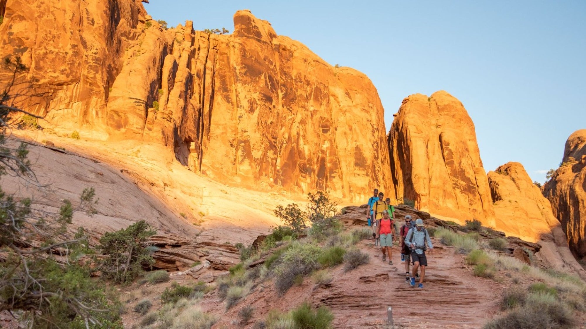 Three hikers walk down a rocky trail surrounded by desert vegetation and large, sunlit red rock formations.