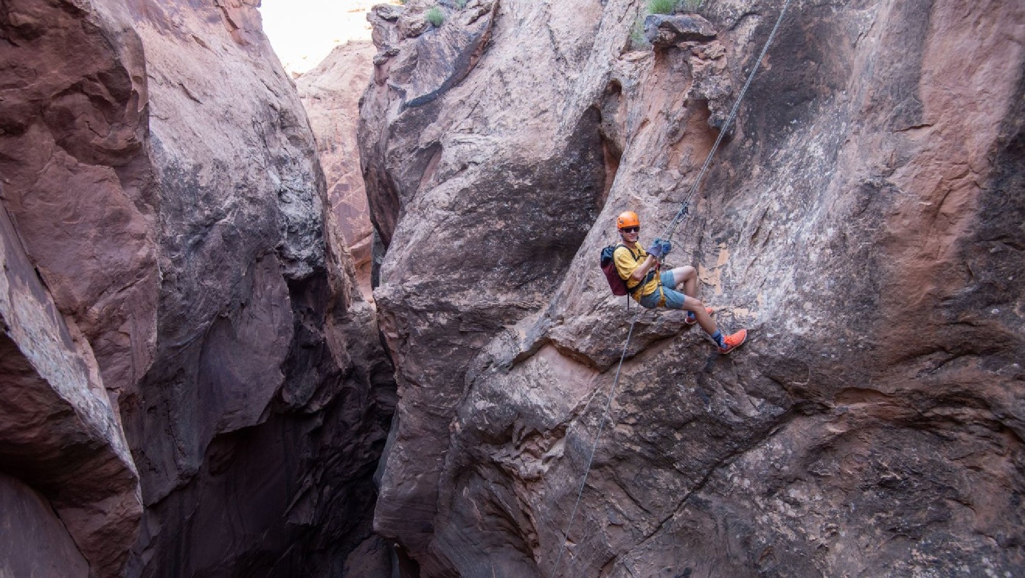 A person wearing a helmet and harness rappels down a steep rock face in a narrow canyon.