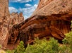 Natural stone arch formation with layered reddish-brown rock, surrounded by green vegetation, under a partly cloudy blue sky.