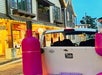 White vehicle with two giant pink bottles on the back parked on a street lined with buildings.