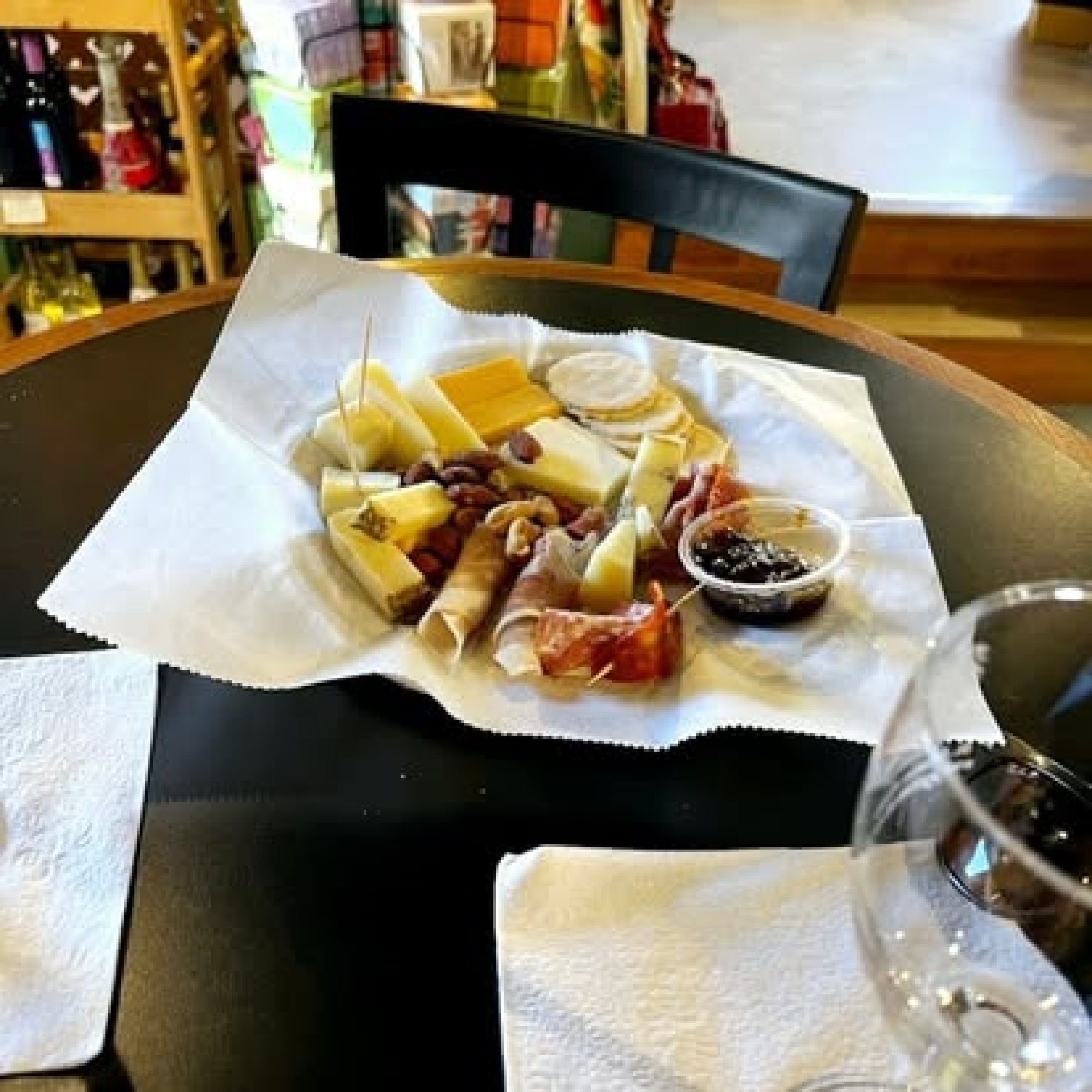 A plate on a table with assorted cheeses, meats, crackers, grapes, and a cup of jam. A wine glass is nearby, and shelves with items are visible in the background.