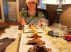 A woman sits at a table with plates of assorted desserts and glasses of wine in front of her. She is wearing a pink cap and a camouflage shirt.