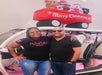 Two people smiling in front of a decorated golf cart with Christmas plush figures and a "Merry Christmas" banner on top. The woman wears a "wine season" shirt, the man wears sunglasses.