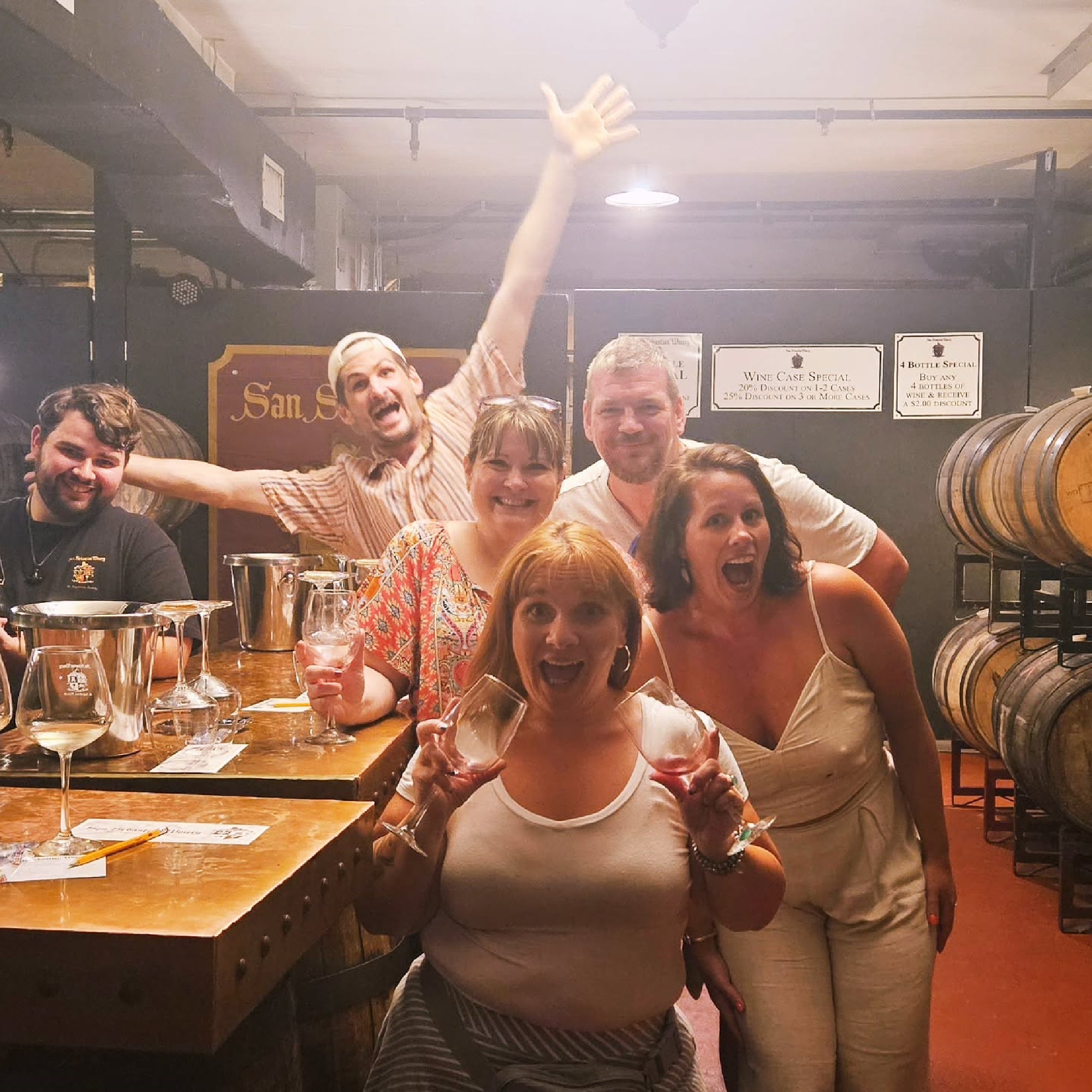 A group of six people enjoy a winery tour, holding wine glasses and posing energetically in front of wooden barrels.