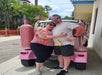 A couple poses in front of a parked pink vehicle decorated with giant wine bottle props on a sunny street.