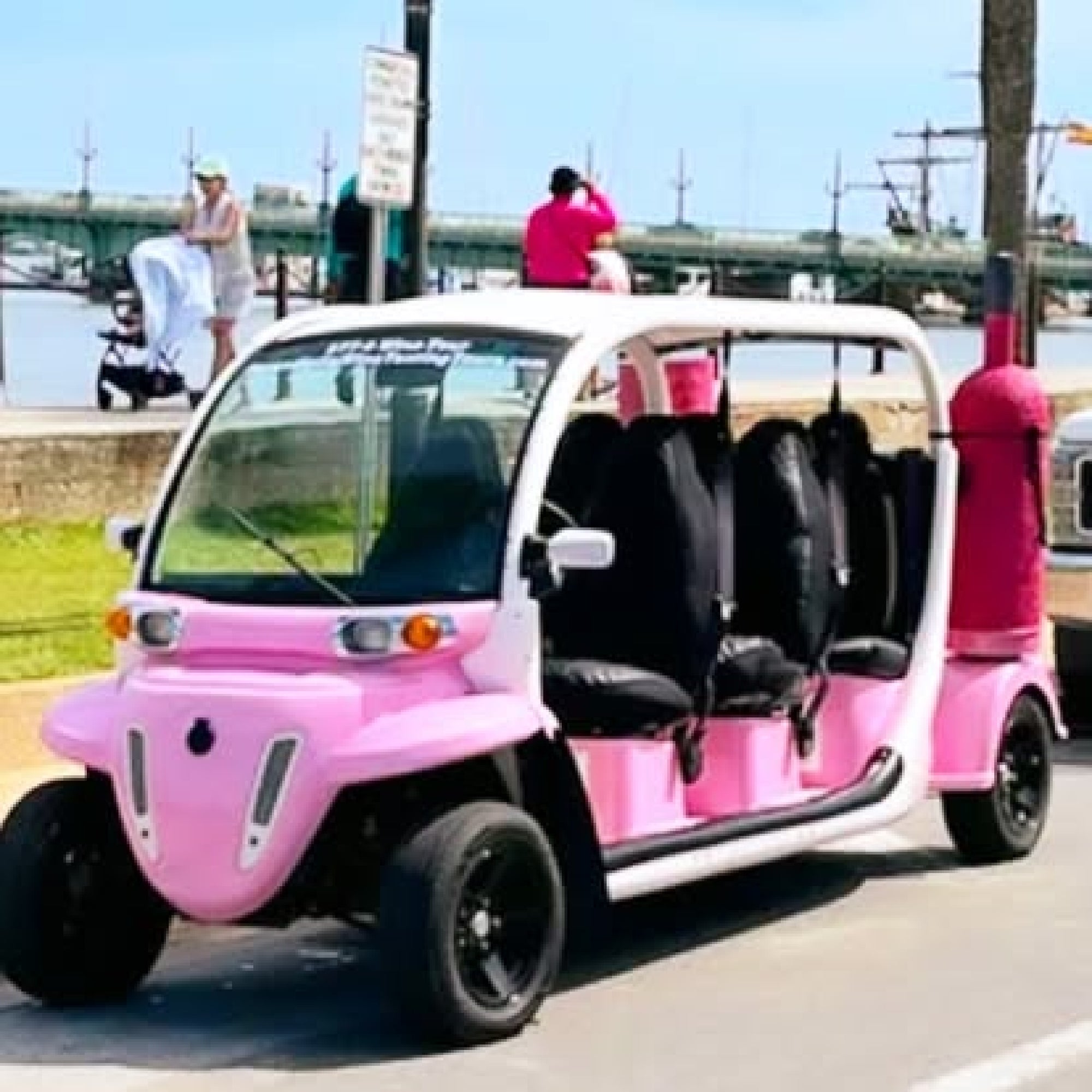 A pink and white, open-sided electric golf cart is parked on a sunny street near a waterfront, with a bridge and ships in the background.