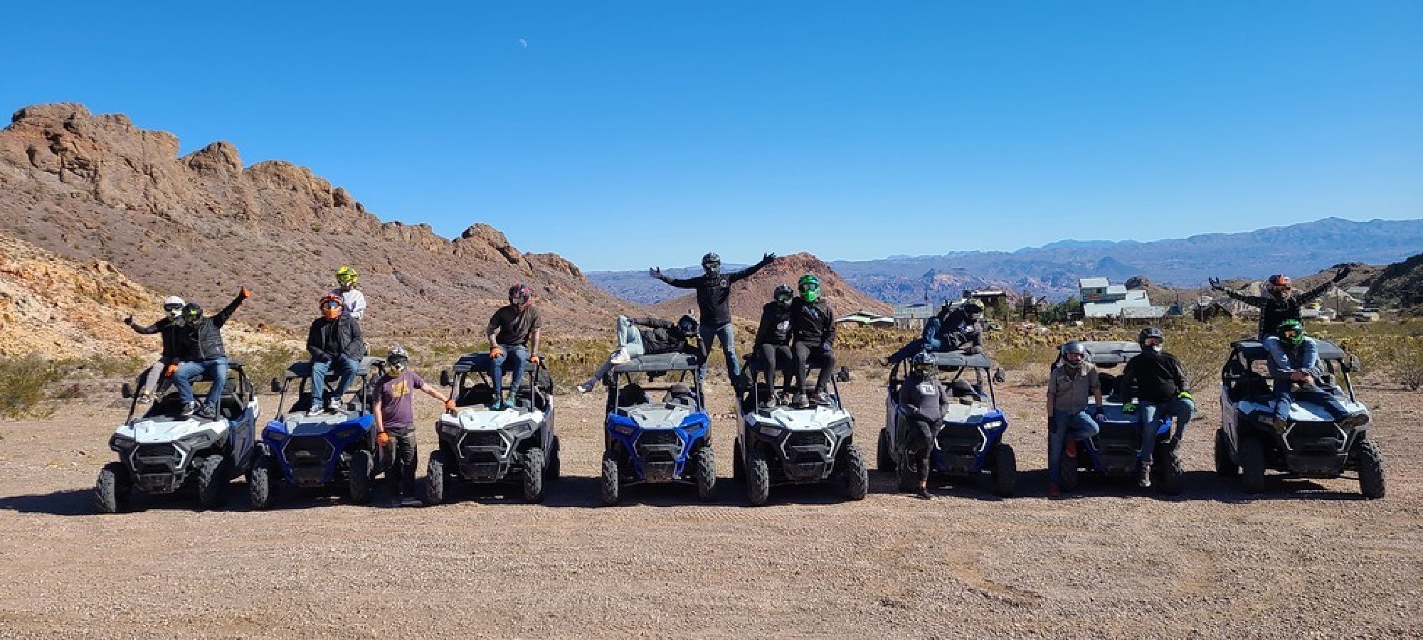 A group of people posing on and next to all-terrain vehicles parked side by side in a desert landscape with rocky hills in the background.