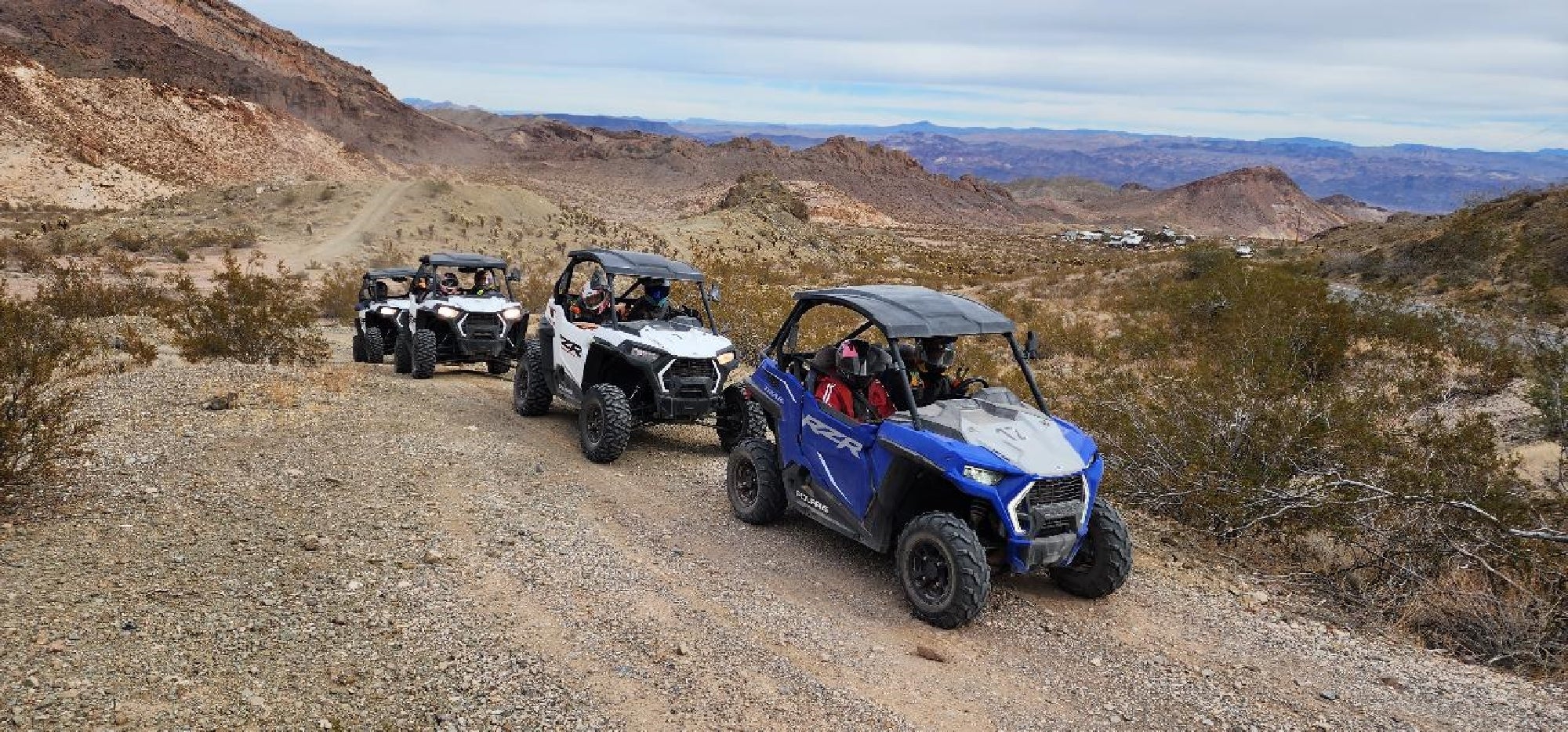 Four off-road utility vehicles drive on a rocky desert trail surrounded by mountains and sparse vegetation under a cloudy sky.