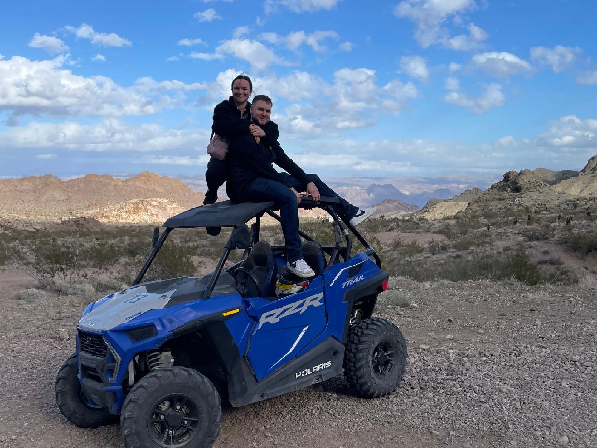 Two people sit on top of a blue Polaris RZR off-road vehicle parked on a rocky desert trail, with mountains and a partly cloudy sky in the background.