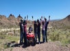 Six people wearing helmets stand with their arms raised beside a red ATV in a desert landscape with mountains in the background under a clear blue sky.