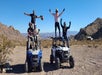Five people wearing helmets pose on two off-road vehicles in a desert landscape with mountains in the background under a clear blue sky.