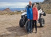 Two people stand in front of an off-road vehicle in a rocky desert area with mountains and scattered buildings in the background.