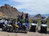 A group of people wearing helmets and gear sit on ATVs and off-road vehicles in a desert landscape with mountains in the background.