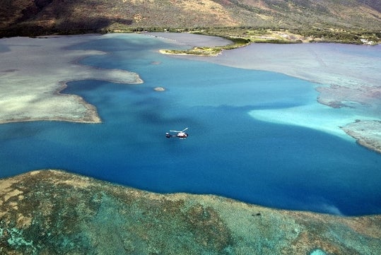 Aerial view looking down on the Molokai Voyage Helicopter Tour over Hawaii's Molokai and Maui islands with bright blue water between them.