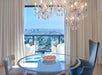 Dining room with a round table and chairs in front of a large window. Chandelier overhead and a bowl of oranges on the table. The window offers a cityscape view with clear skies.