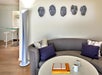 Modern living room with a curved sofa, purple cushions, a round table with books and mugs, wall-mounted face sculptures, and a tall lamp.