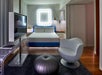 Modern hotel room with a double bed, a large TV, a glass desk, a round gray chair, and a circular ottoman. Lamps hang on either side of the bed, and a lighted frame is on the wall.