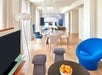Modern living room with wooden flooring, blue and white chairs, a circular dining table, two gray stools, a black round coffee table with oranges, a TV, and two chandeliers.