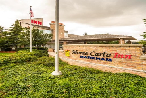Entrance of Monte Carlo Inn Markham, featuring a brick sign and a U.S. flag on a pole, with trees and a cloudy sky in the background.