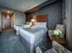 Hotel room with two double beds, patterned bedding, a lamp on a shared nightstand, two chairs with a small round table, and a vase of flowers. Walls are covered with dark textured wallpaper.