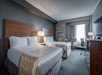 Hotel room with two double beds, a large window, a TV, an armchair, and a small table. Neutral-toned decor with wooden accents and a patterned carpet.