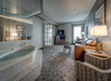 Hotel room with a large bed, armchair, TV, and a jacuzzi tub. The room features patterned carpet, a window with curtains, and decorative lighting.