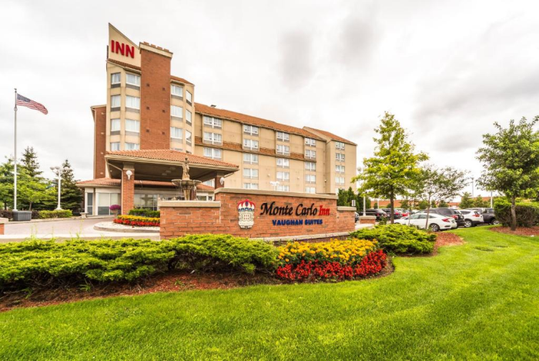 A hotel with a sign reading "Monte Carlo Inn Vaughan Suites" features a landscaped entrance and parking lot. An American flag is visible.