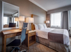 Hotel room with a large bed, wooden desk, office chair, and two lamps. Two windows with curtains allow natural light to fill the room.