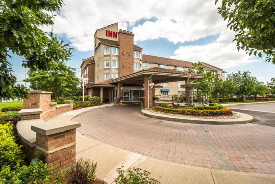 Front view of a hotel labeled "INN" with a covered entrance, circular driveway, and landscaped gardens under a partly cloudy sky.