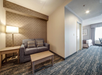 Hotel room with a sitting area featuring a sofa, lamp, and table; a bed is visible through an open doorway, with patterned carpet throughout.